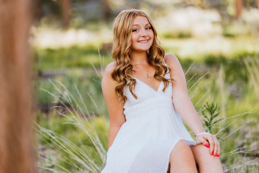 Young woman in white dress sitting outdoors among tall grass, smiling gracefully in a sunlit natural setting.