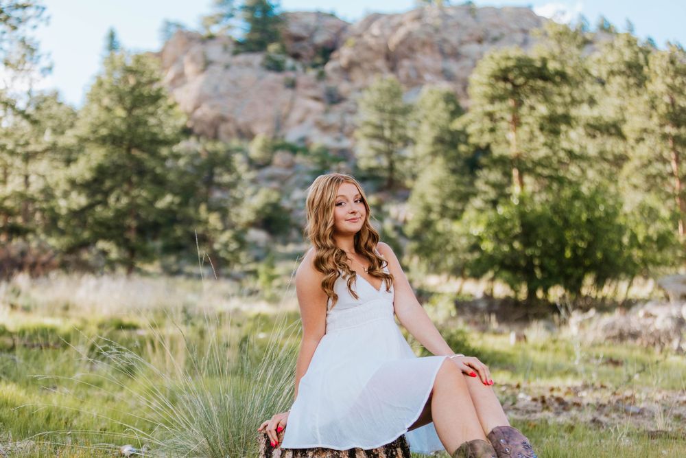 Young woman in white dress and boots sitting outdoors with rocky mountain landscape and green trees behind her.