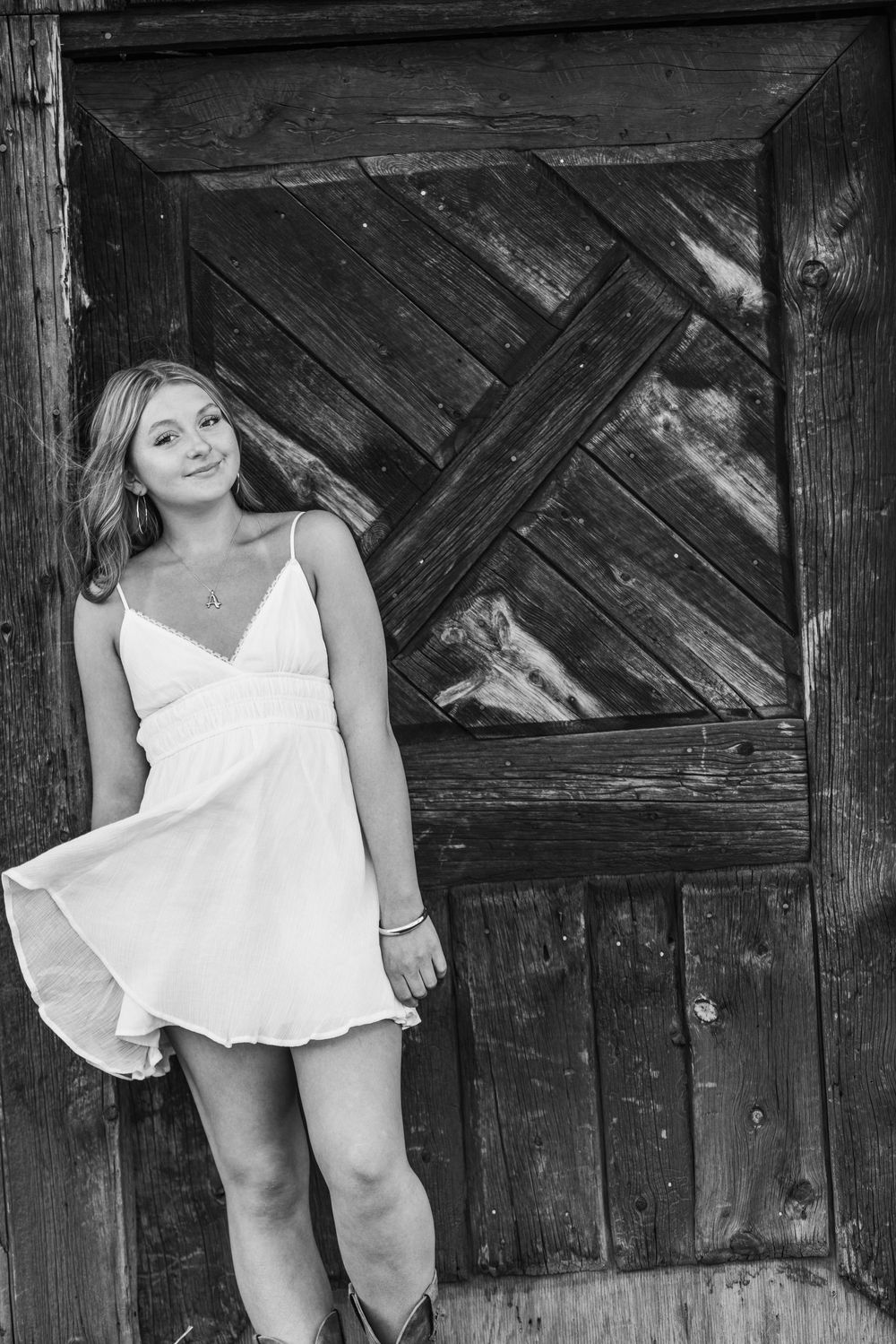 Young woman in white sundress and cowboy boots leaning against rustic wooden barn door, black and white photo.