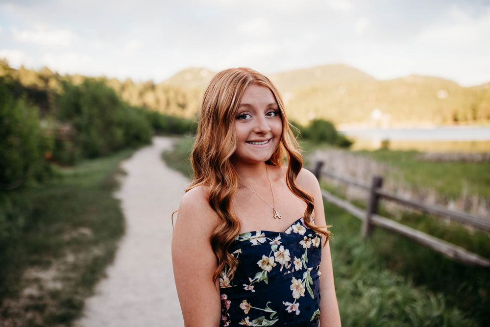 Young woman with long red hair in floral dress smiling on scenic mountain path at golden hour.