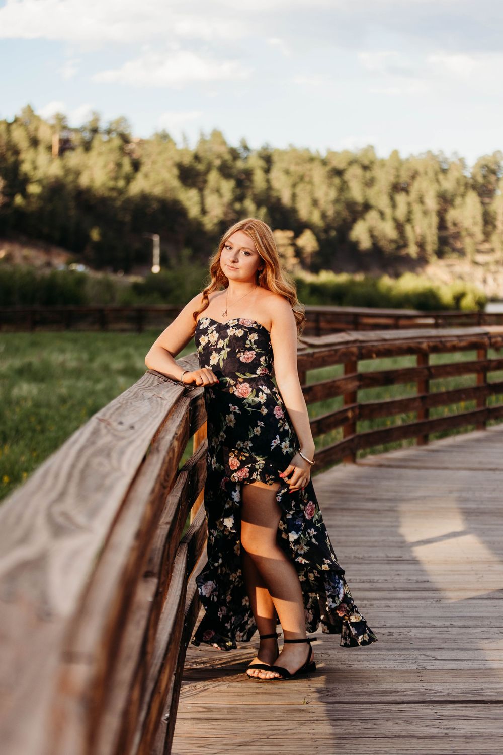 Young woman in black floral maxi dress posing on wooden bridge with lush green forest backdrop.