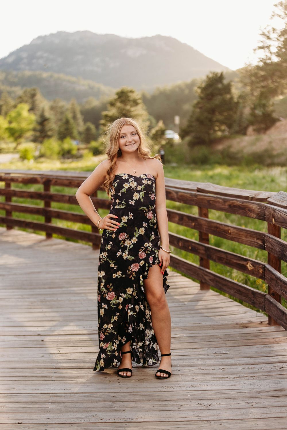 Smiling blonde woman in black floral dress standing on wooden bridge with mountain scenery behind.