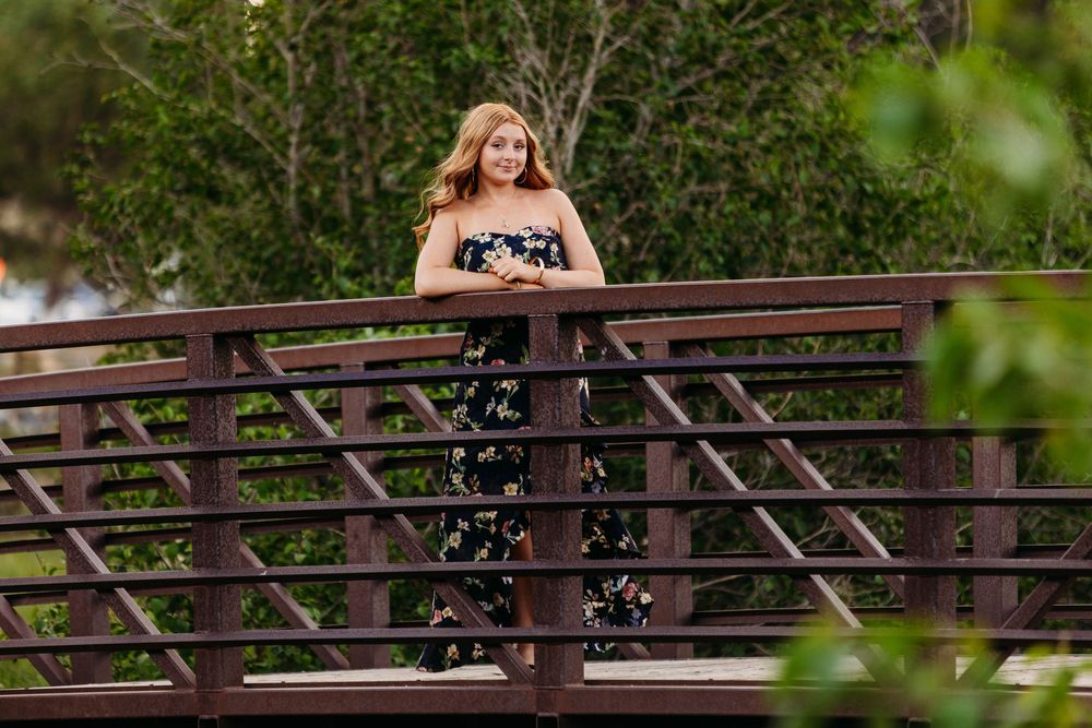 Young woman in floral dress posing on a wooden bridge surrounded by lush green trees.