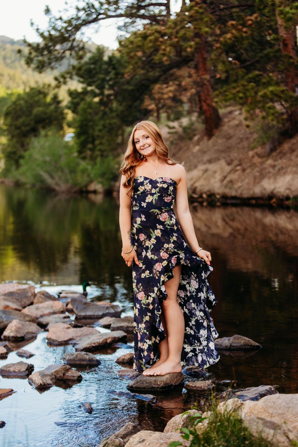 Young woman in floral strapless maxi dress standing on rocks by a serene mountain river surrounded by lush trees.