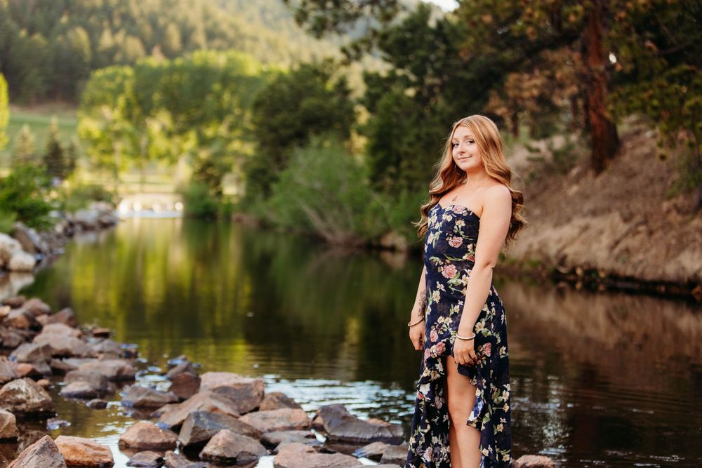 Young woman in floral maxi dress standing by a rocky river surrounded by lush green trees in golden hour light.