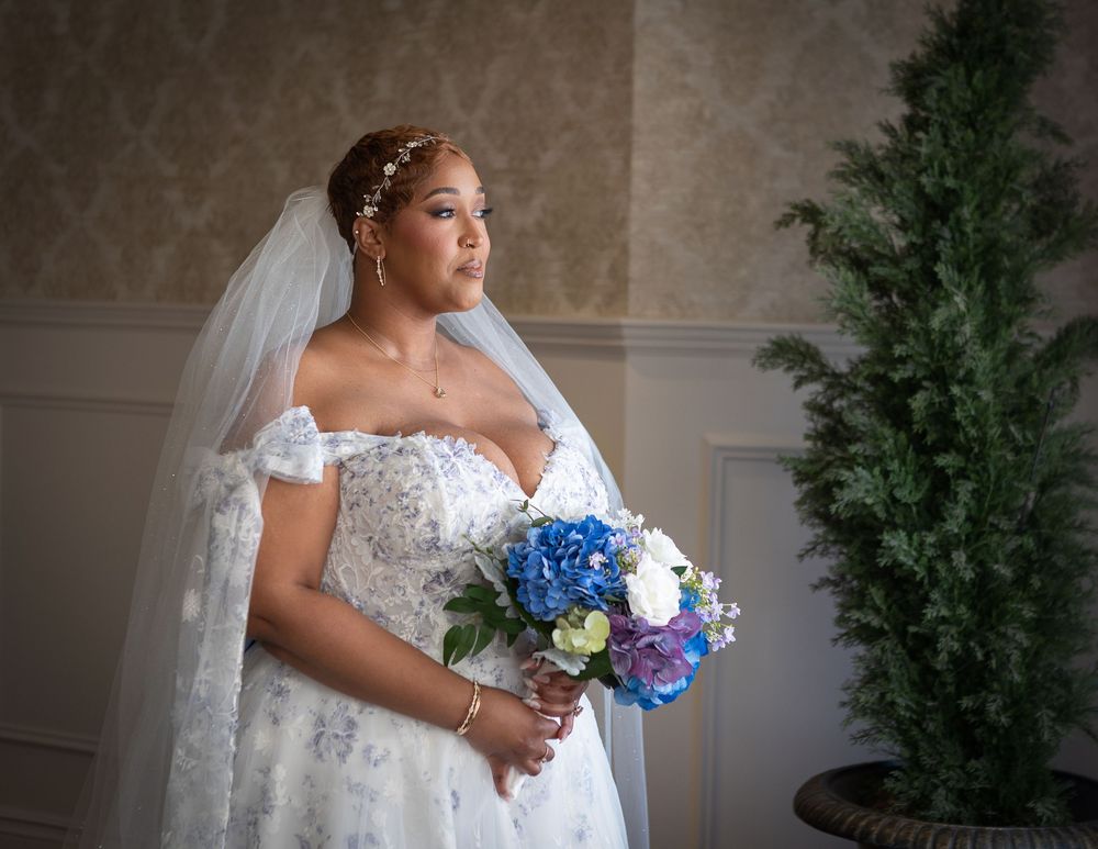Elegant bride in lace off-shoulder gown holding blue hydrangea bouquet, wearing veil with pearl headpiece indoors.