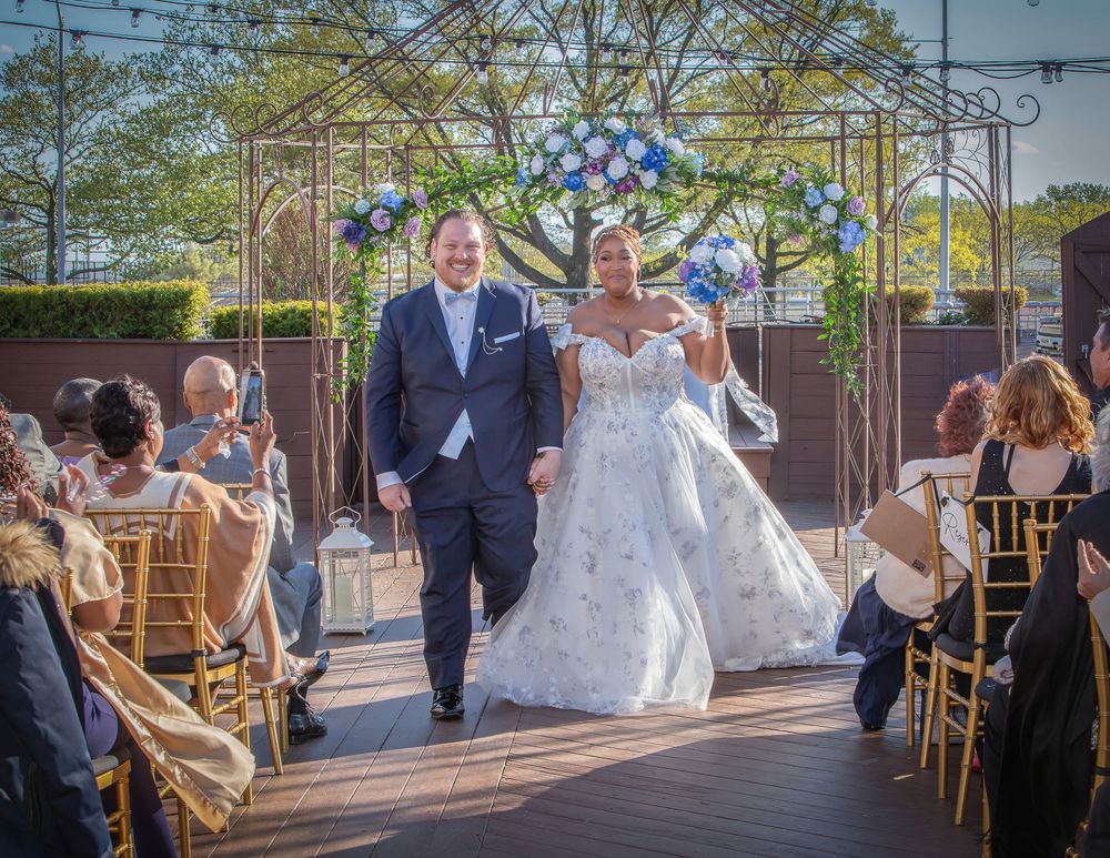 Bride and groom walk down the aisle at an outdoor wedding ceremony with blue floral arch decor.