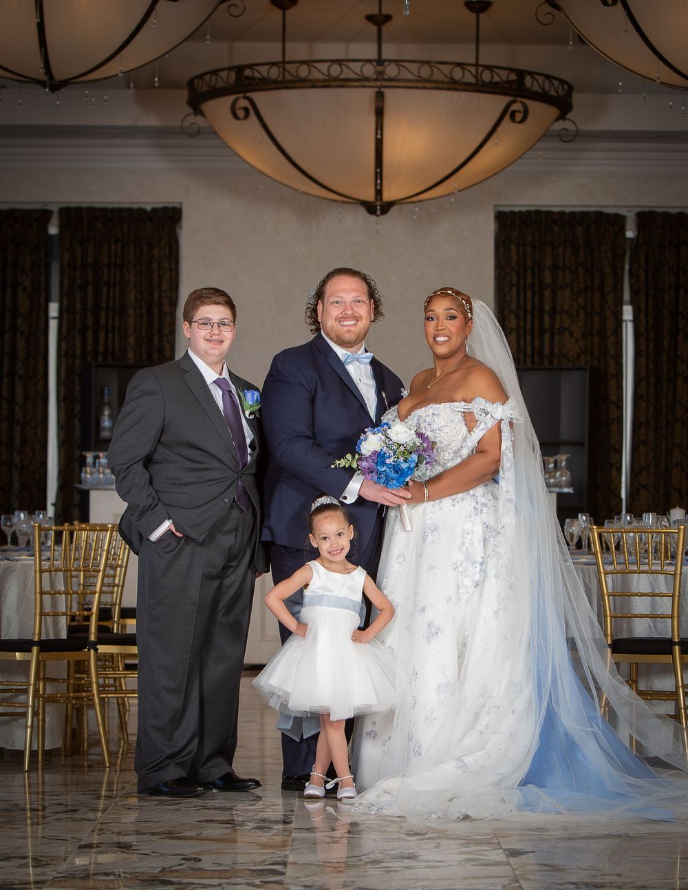 Bride and groom pose with two children in elegant wedding venue, bride in lace gown holding blue bouquet.