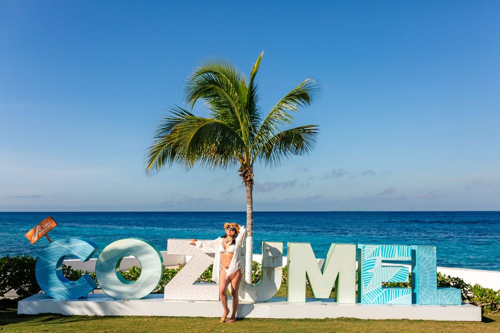 Woman posing by colorful Cozumel sign near palm tree with turquoise Caribbean Sea in background.