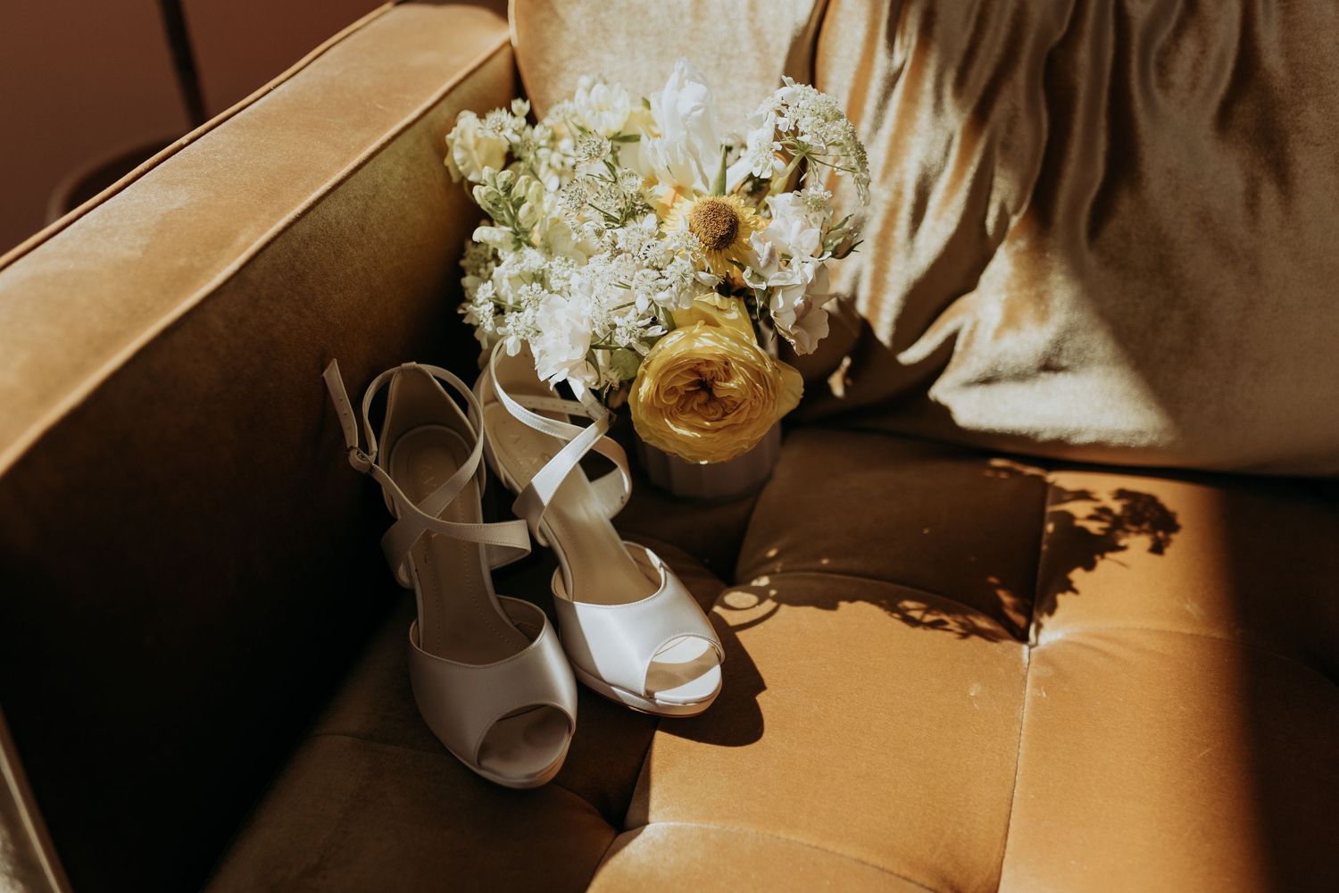 A yellow bouquet of flowers and white wedding shoes on a velvet chair in the sunlight.