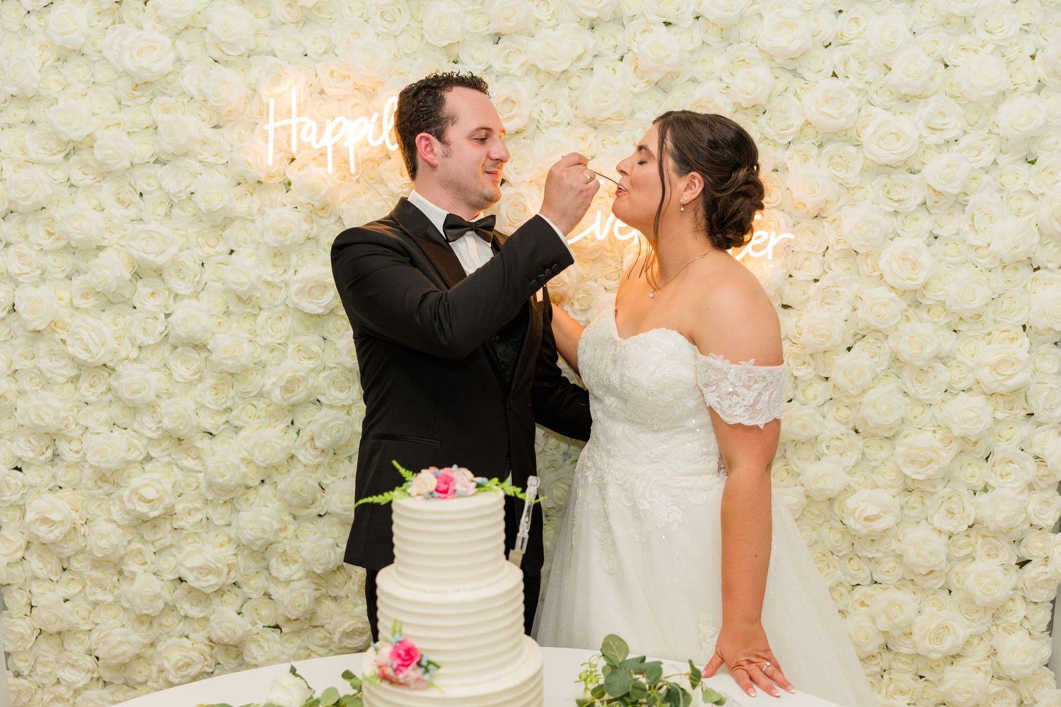 Bride and groom feeding each other wedding cake in front of a white flower wall with a neon Happily Ever After sign.
