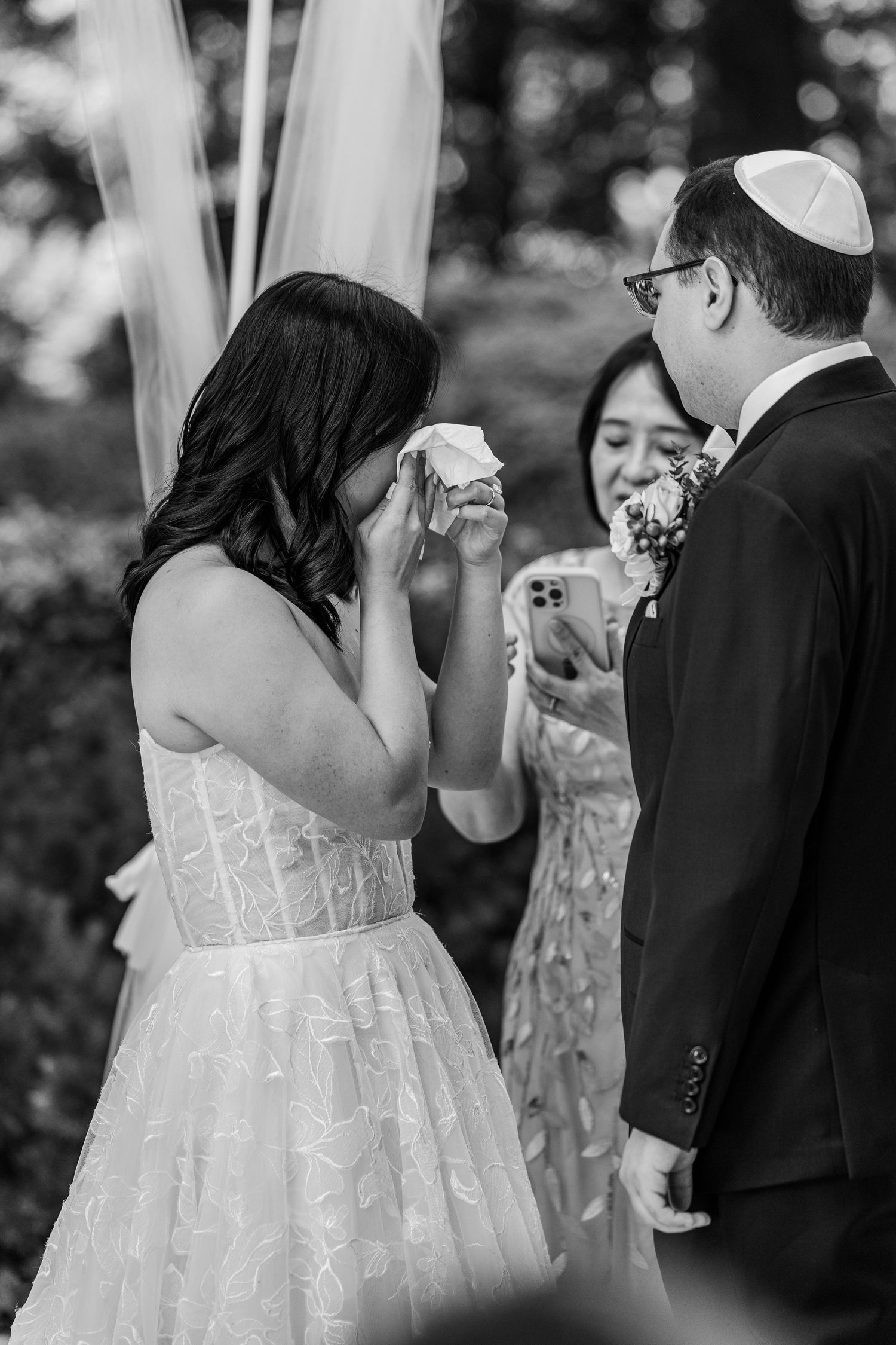 Black and white photo of emotional bride wiping tears during outdoor Jewish wedding ceremony.