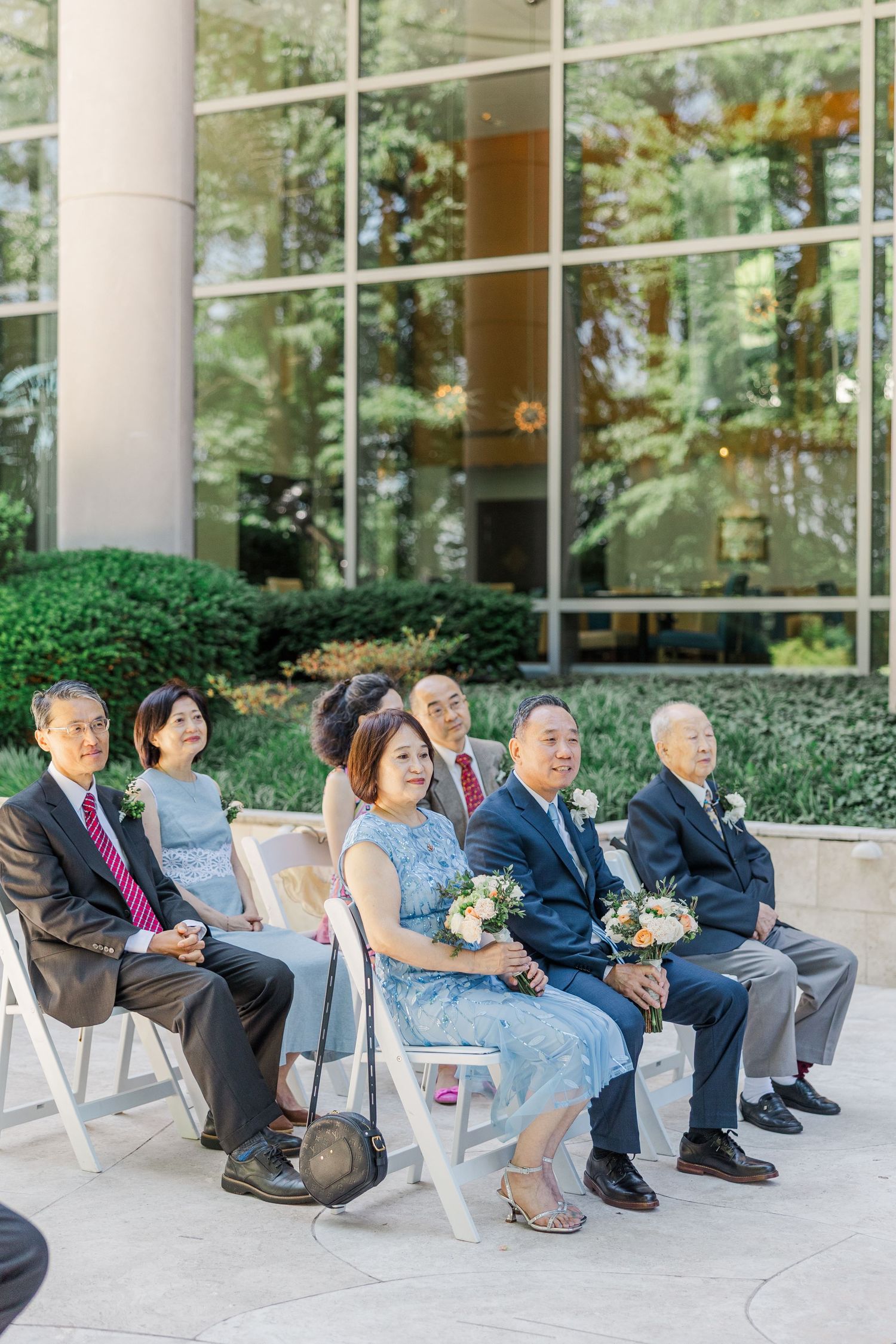 Guests seated in white chairs at an outdoor wedding ceremony, dressed in formal attire, holding bouquets near a glass building.