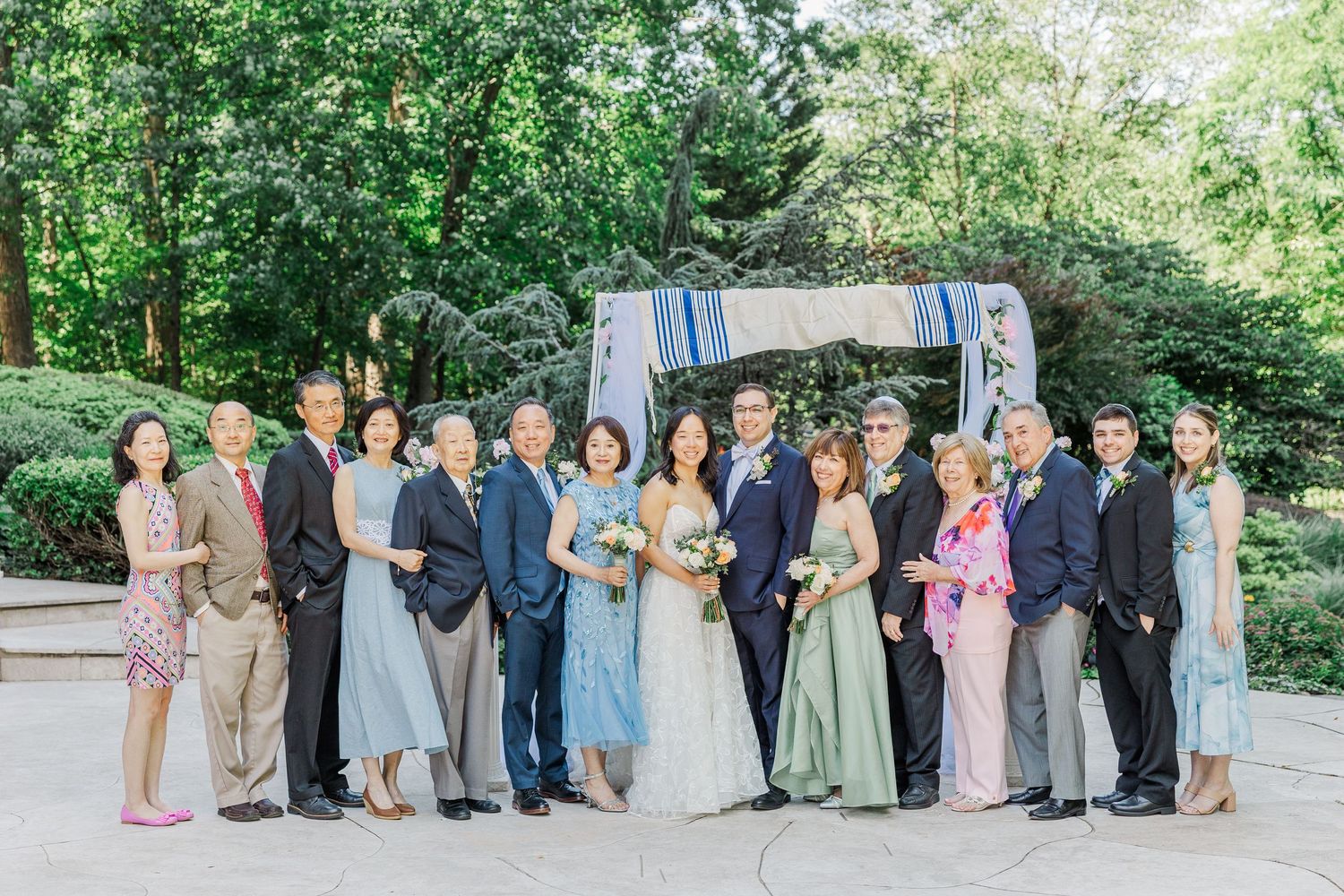A wedding party poses outdoors in formal attire, surrounded by lush greenery, celebrating a joyful occasion.