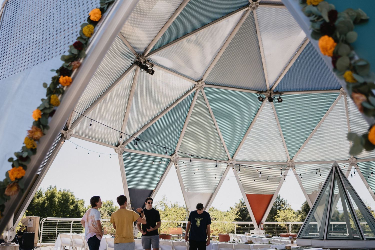 People gather beneath a colorful geometric dome tent decorated with marigold garlands and string lights at an outdoor event.