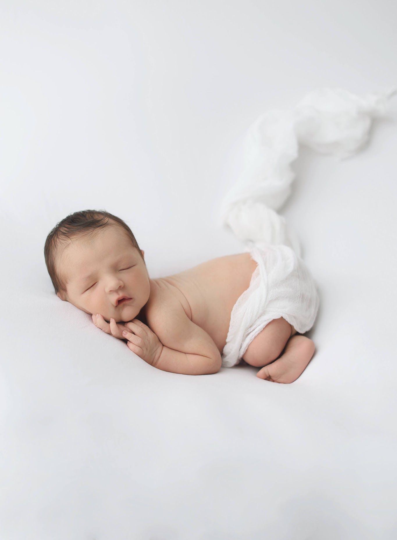 Adorable newborn baby sleeping curled on side with white swaddle wrap on bright white background.