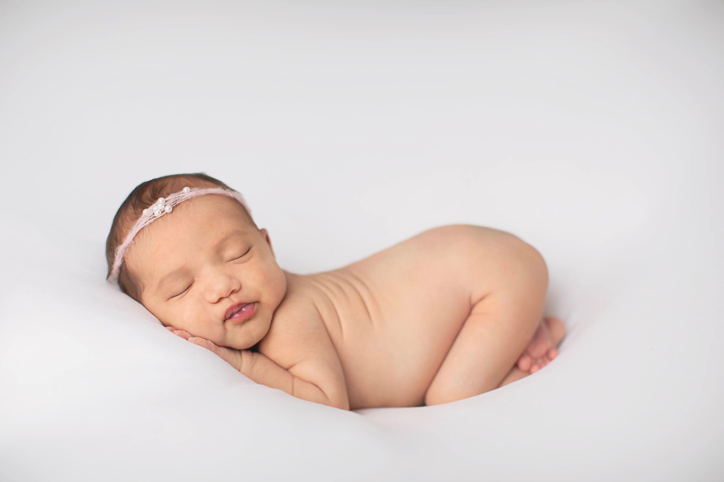 Peaceful newborn baby girl sleeping curled up on white background wearing a delicate floral headband.