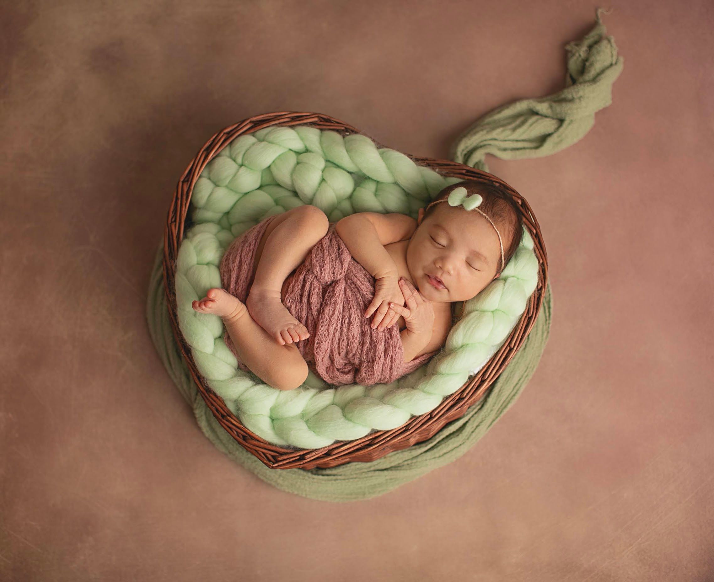 Sleeping newborn baby girl in pink wrap nestled in wicker basket with green chunky knit blanket.