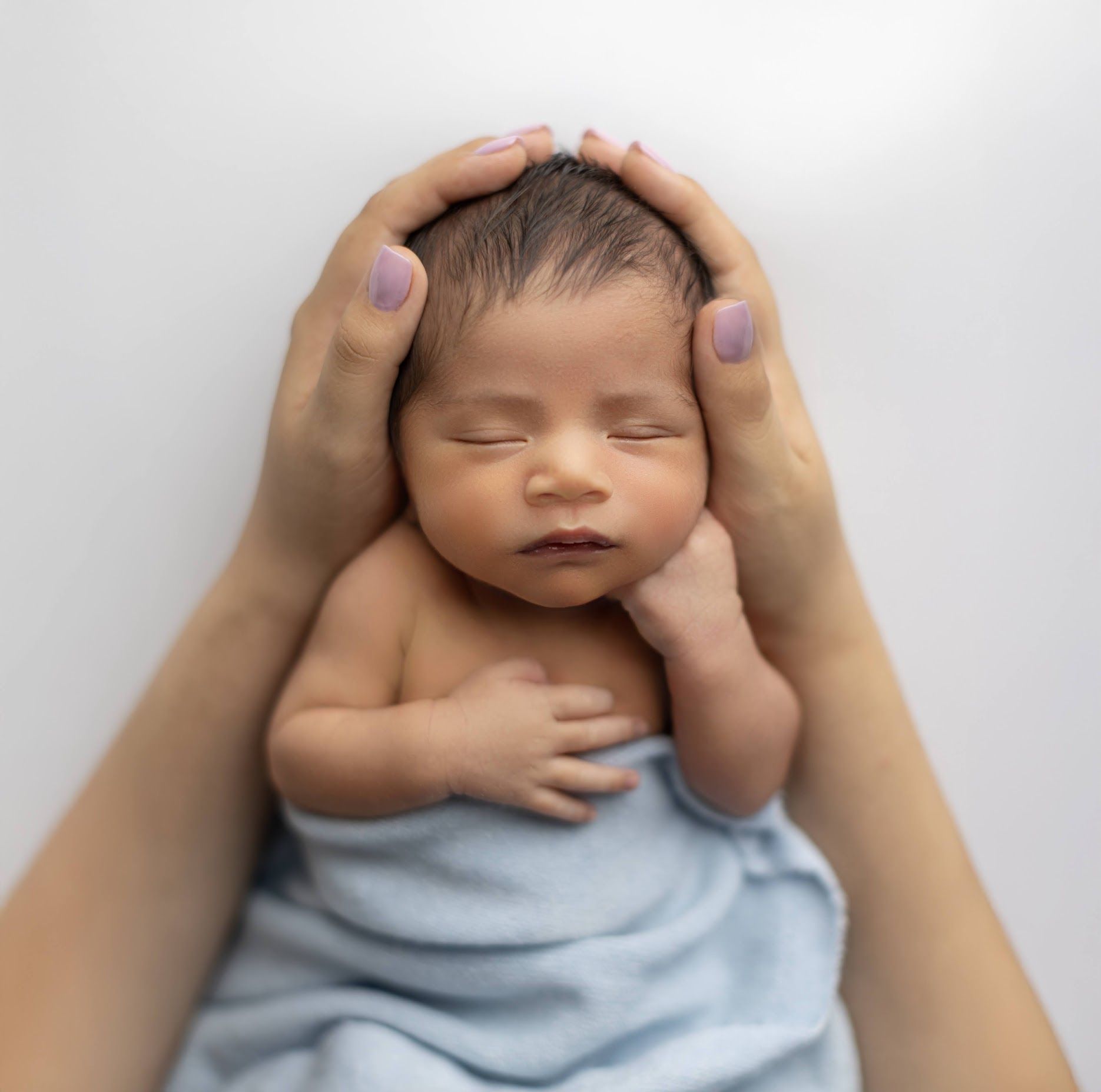 Newborn baby boy wrapped in blue swaddle gently held by mother's hands on white background.