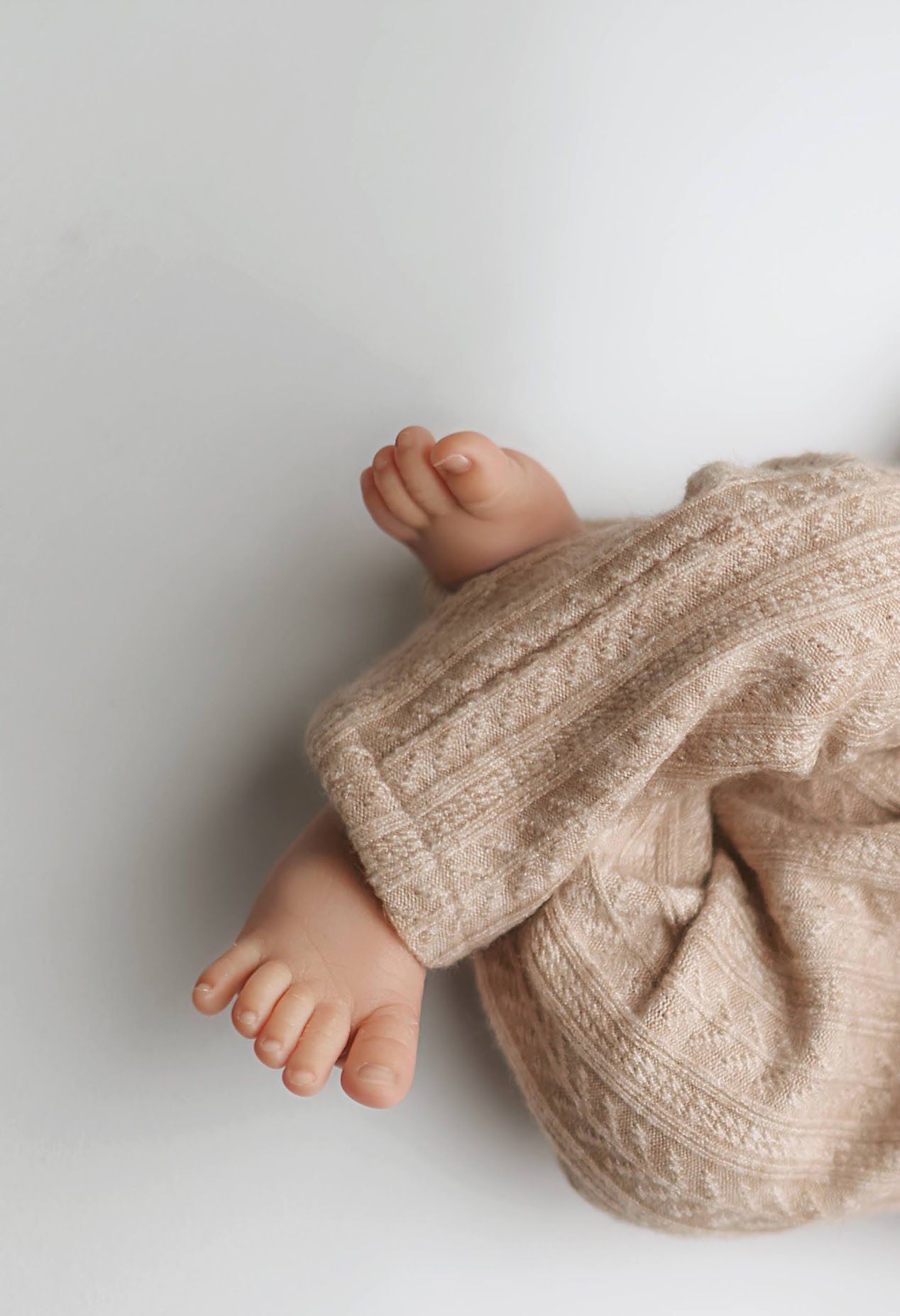 Close-up of newborn baby tiny feet and toes in a soft beige cable-knit outfit on white background.