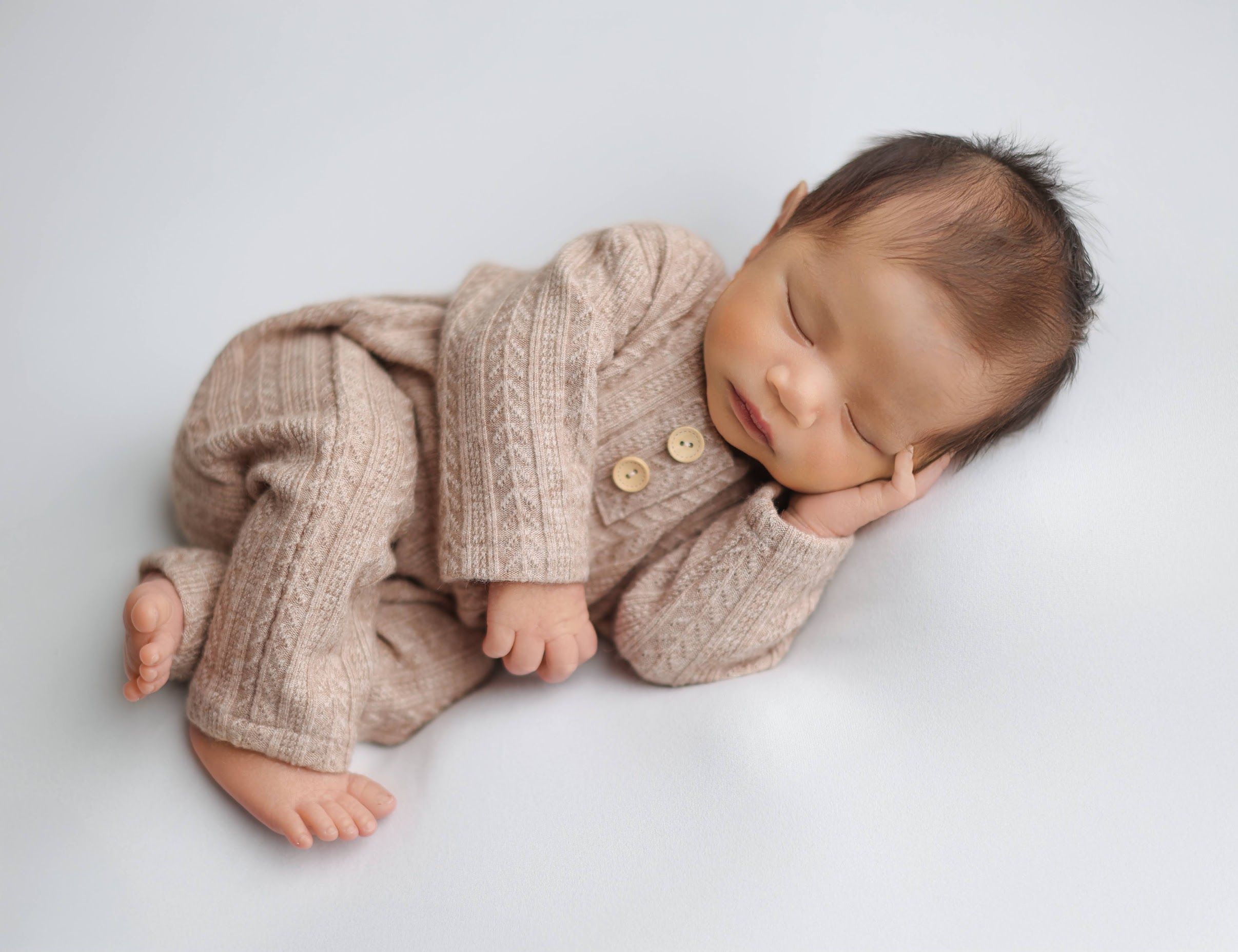 Sleeping newborn baby in beige cable-knit outfit lying on white background with button details.