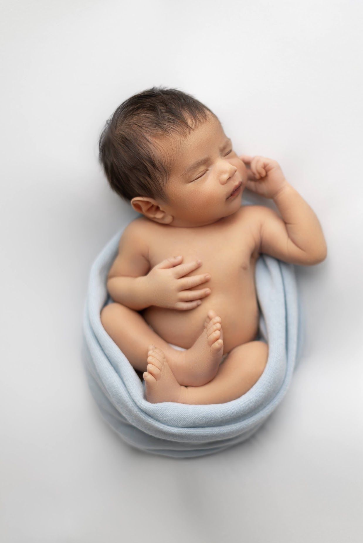 Newborn baby boy sleeping wrapped in light blue swaddle on white background in fetal position.