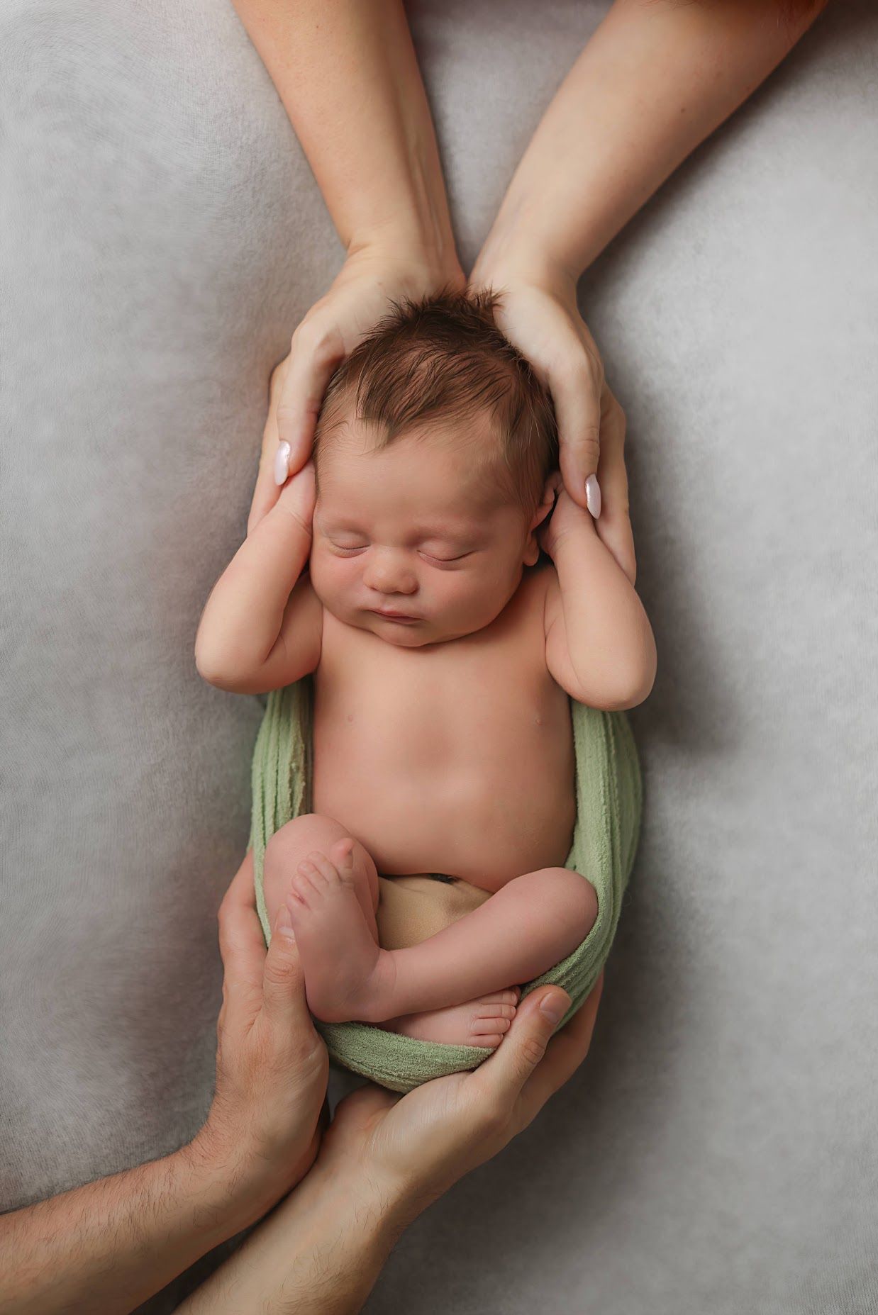 Newborn baby in green wrap cradled by both parents' hands on soft gray background.