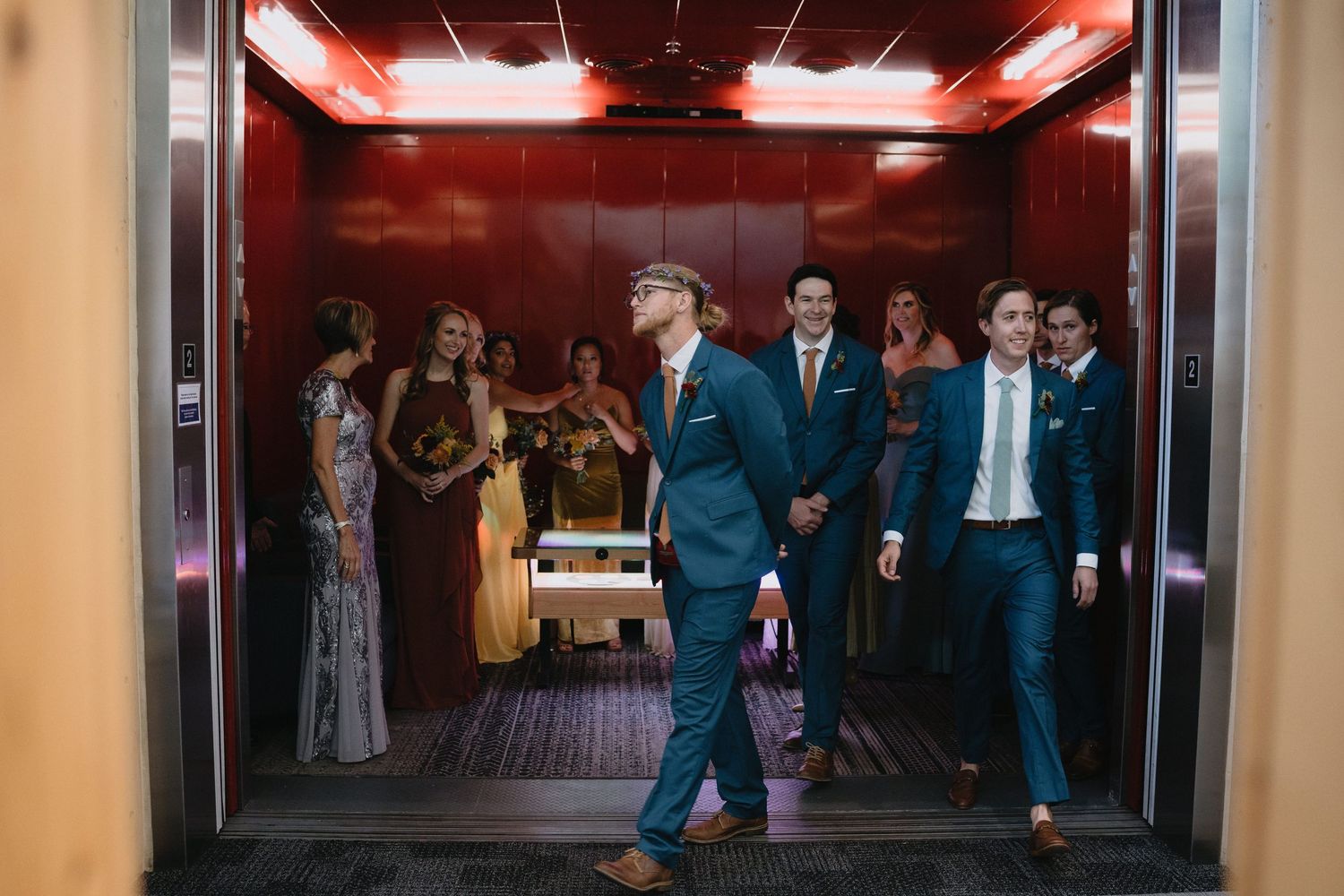 Two grooms in navy suits exit an elevator surrounded by wedding guests in colorful formal attire.