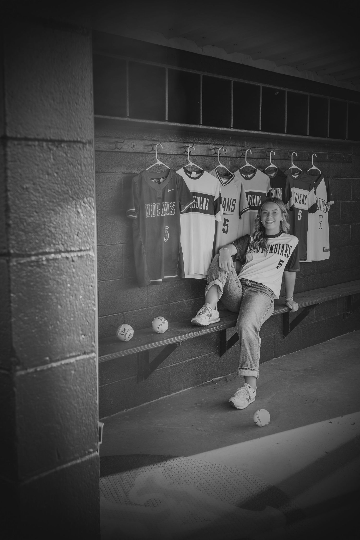 Black and white photo of a softball player sitting in a dugout surrounded by jerseys and softballs.