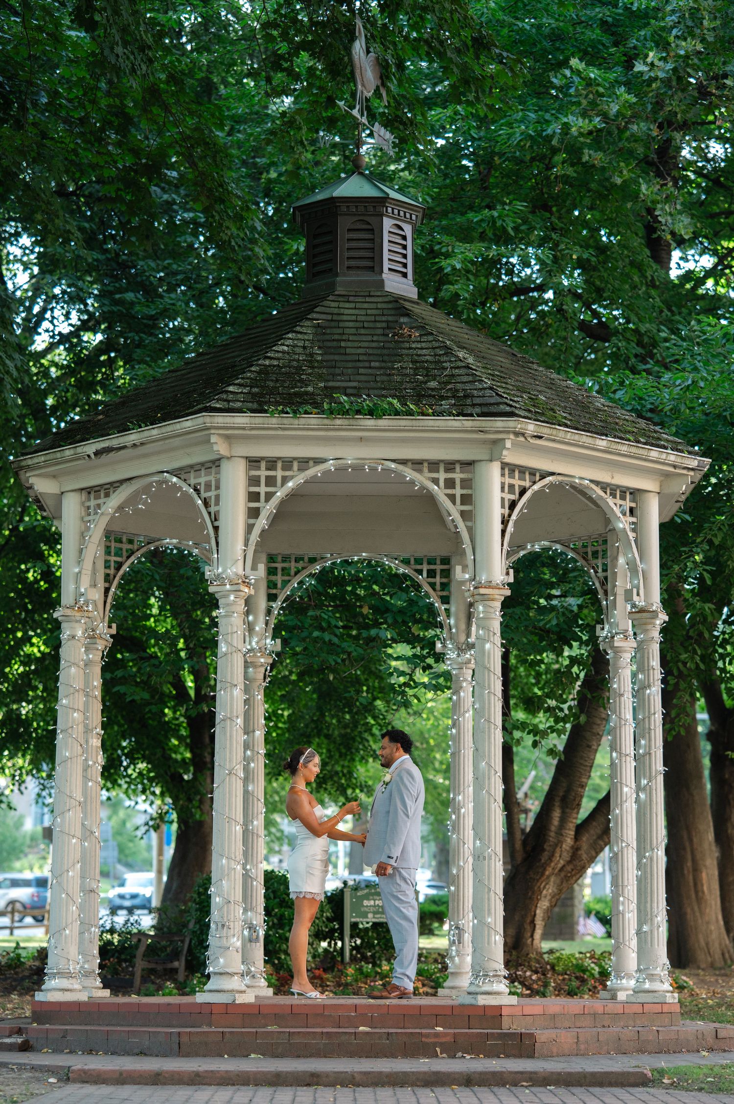 Couple sharing their vows during an elopement in Westchester in a white gazebo decorated with fairy lights, surrounded by lush green trees.