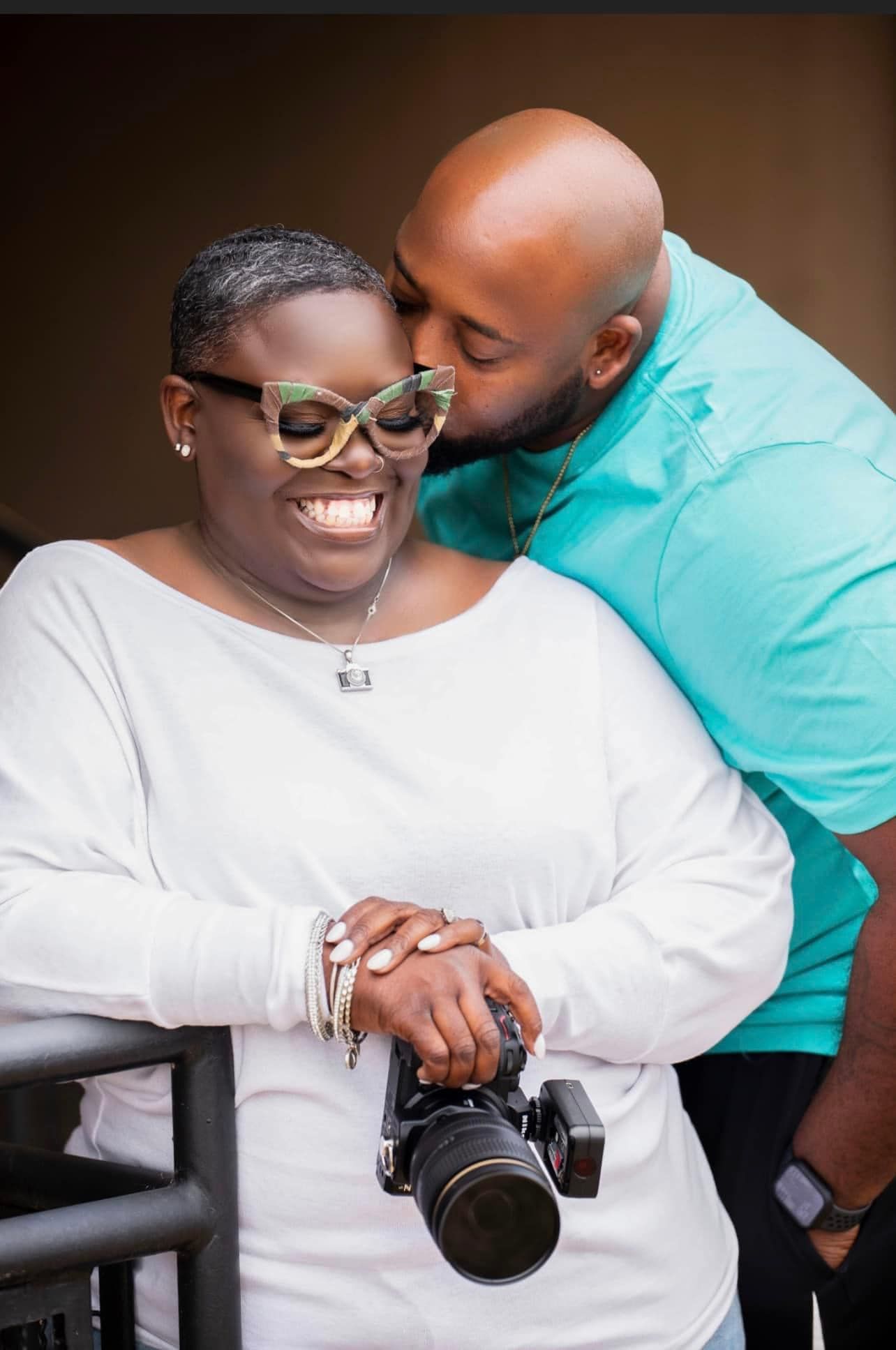 A smiling woman holding a camera receives a kiss on the cheek from a bald man in a teal shirt.