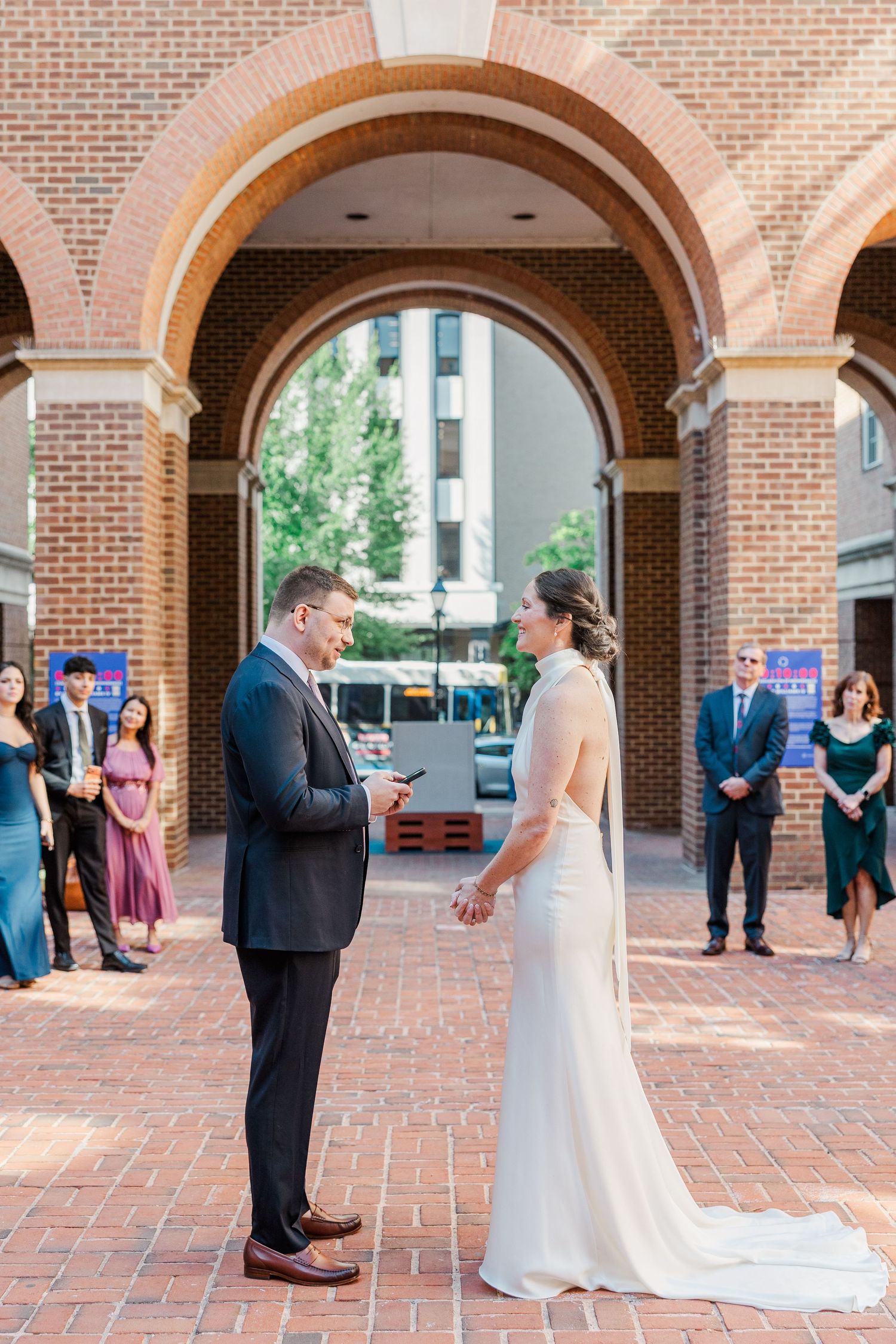 Couple exchanges vows under brick archway at outdoor wedding ceremony with guests watching in background.