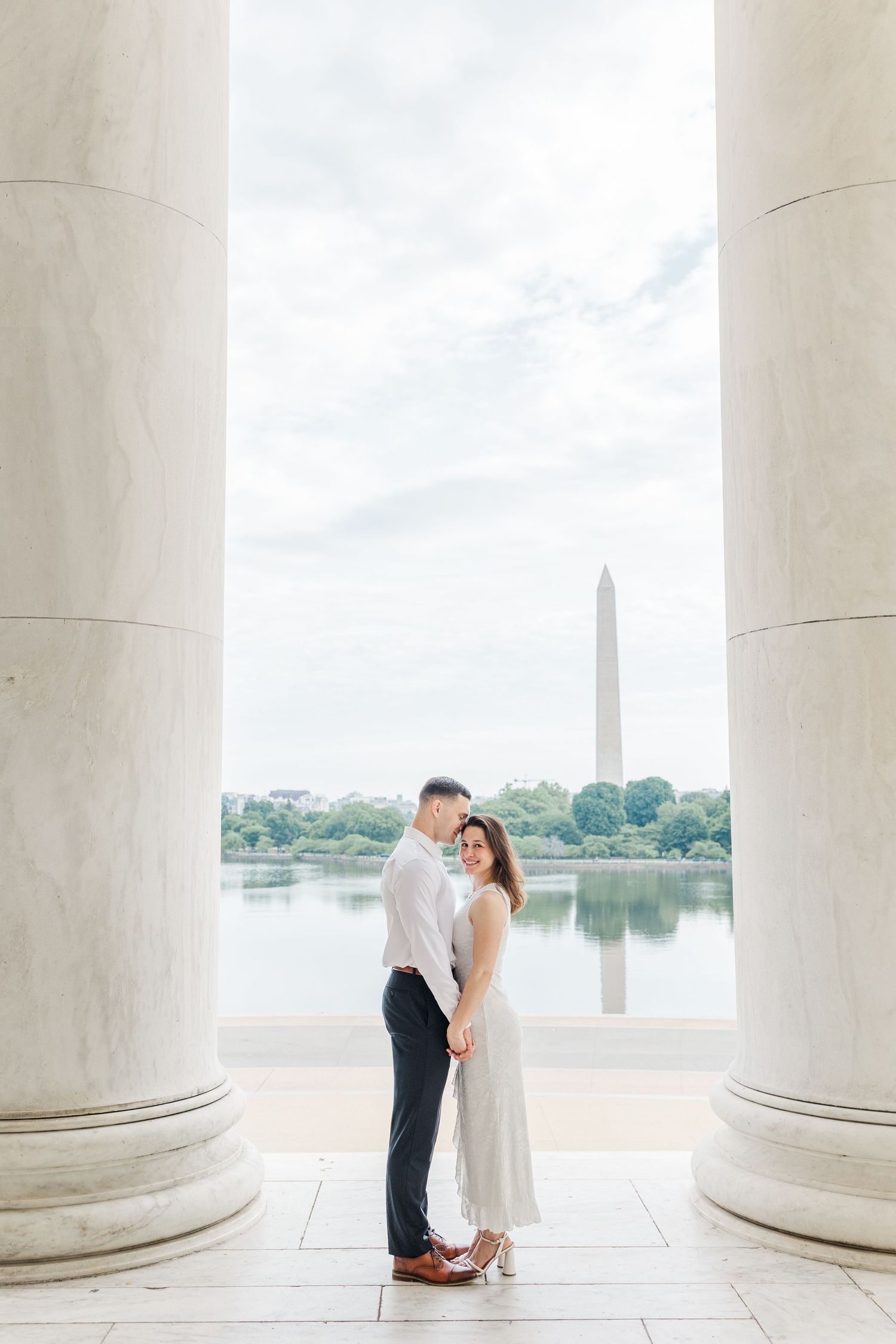 Couple holds hands between marble columns at Jefferson Memorial with Washington Monument reflected in Tidal Basin behind them.