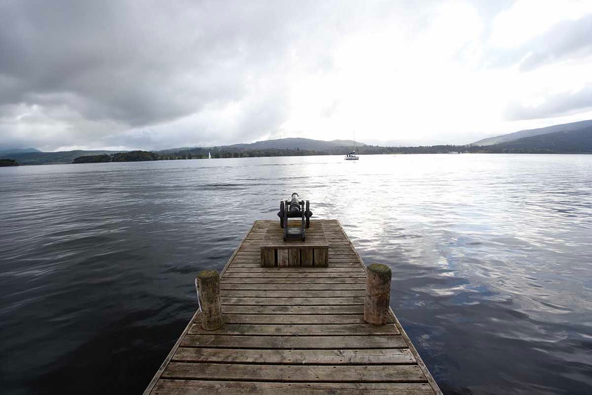 Wooden dock extending over calm lake waters with a chair at the end, mountains and cloudy sky in the background.