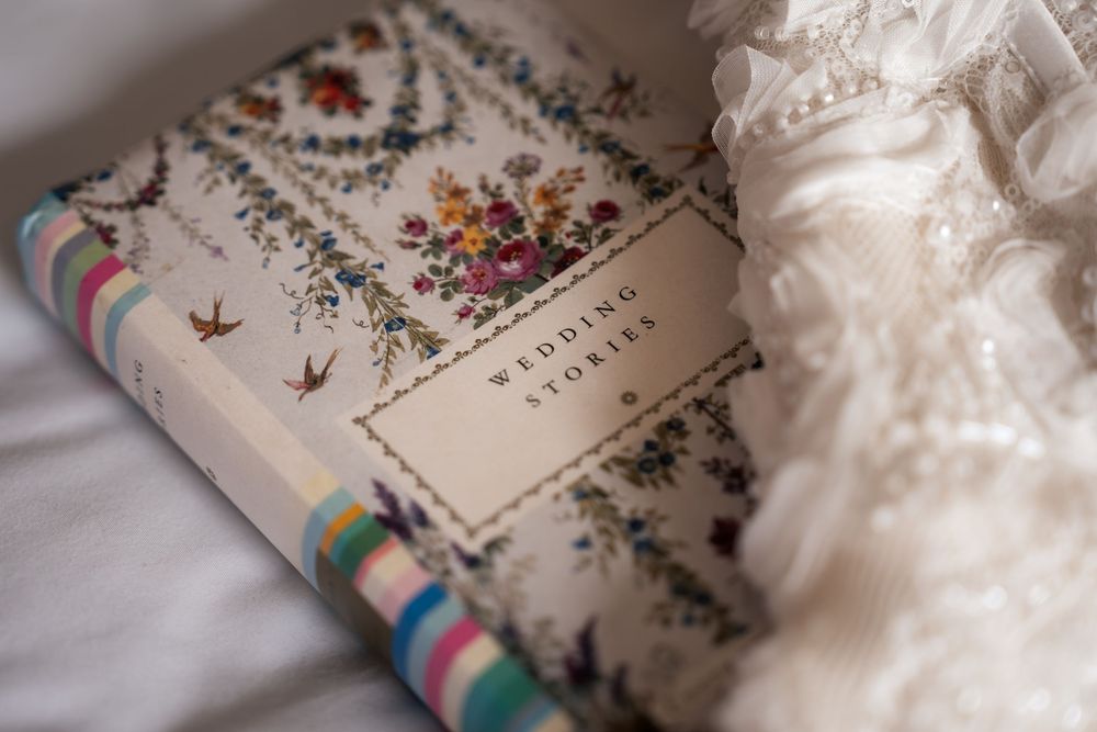 A floral-covered Wedding Stories book rests beside a delicate lace wedding dress on a white surface.