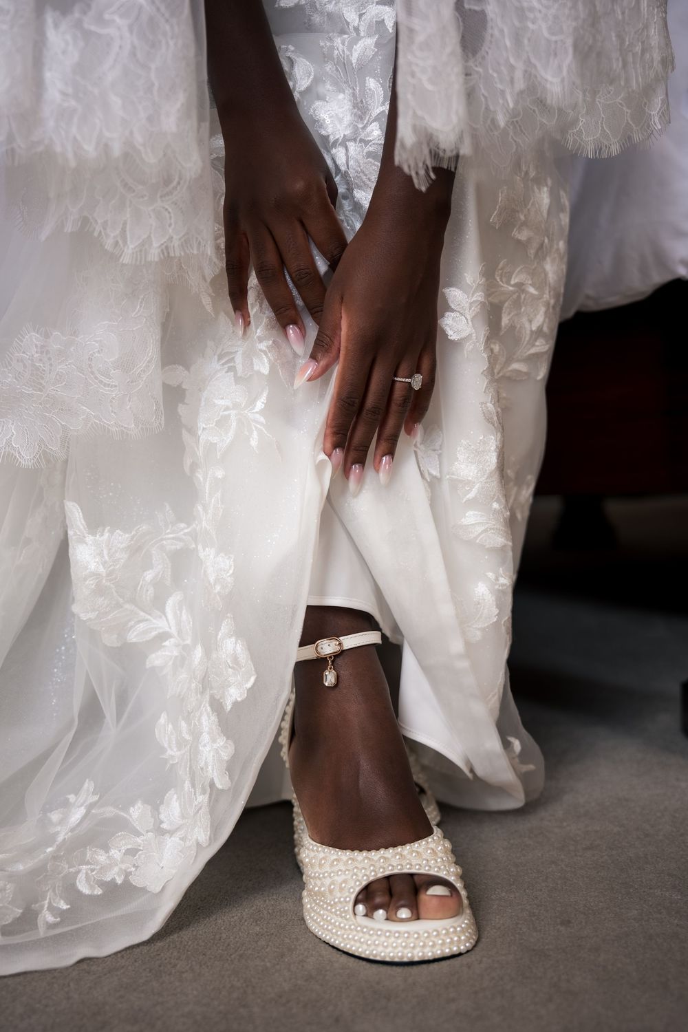 Bride in white lace gown adjusting pearl platform wedding sandal, diamond ring visible on her hand.