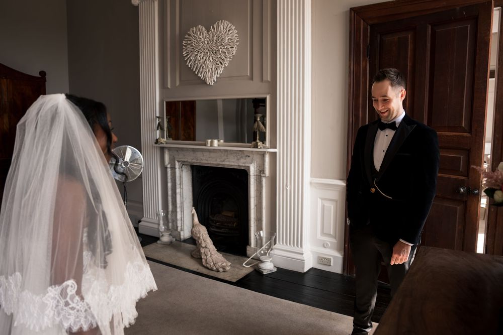 Groom in black tuxedo smiling at bride in white wedding dress and veil, standing by elegant fireplace with heart decor.