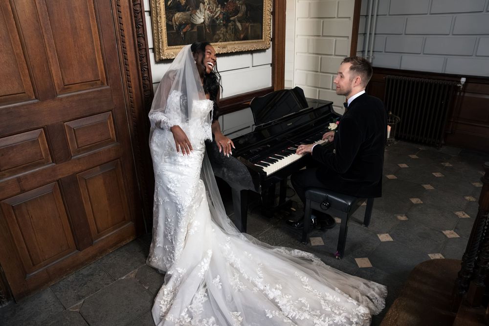 Bride in lace gown with veil stands by groom playing piano at elegant wedding venue.
