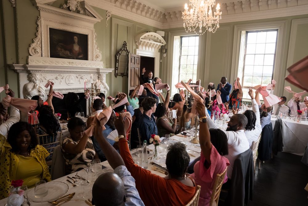 Guests raise their hands in celebration at an elegant wedding reception dinner in a grand Georgian dining room.