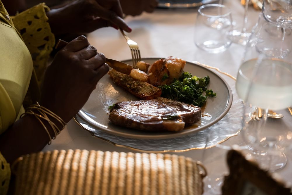 Person cutting a grilled steak with roasted potatoes and greens on a silver plate at an elegant dinner table.