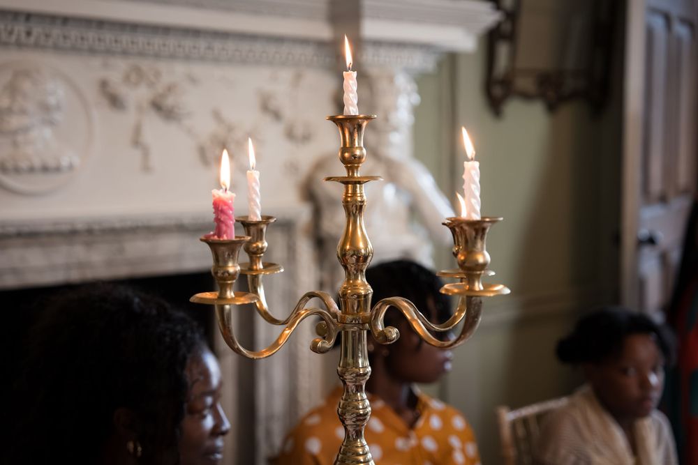A brass candelabra with lit candles stands elegantly in a room with people gathered near an ornate fireplace.