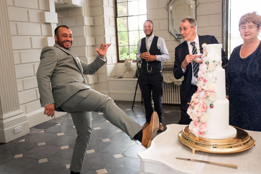 Groom playfully kicks near wedding cake while guests laugh at elegant reception venue.