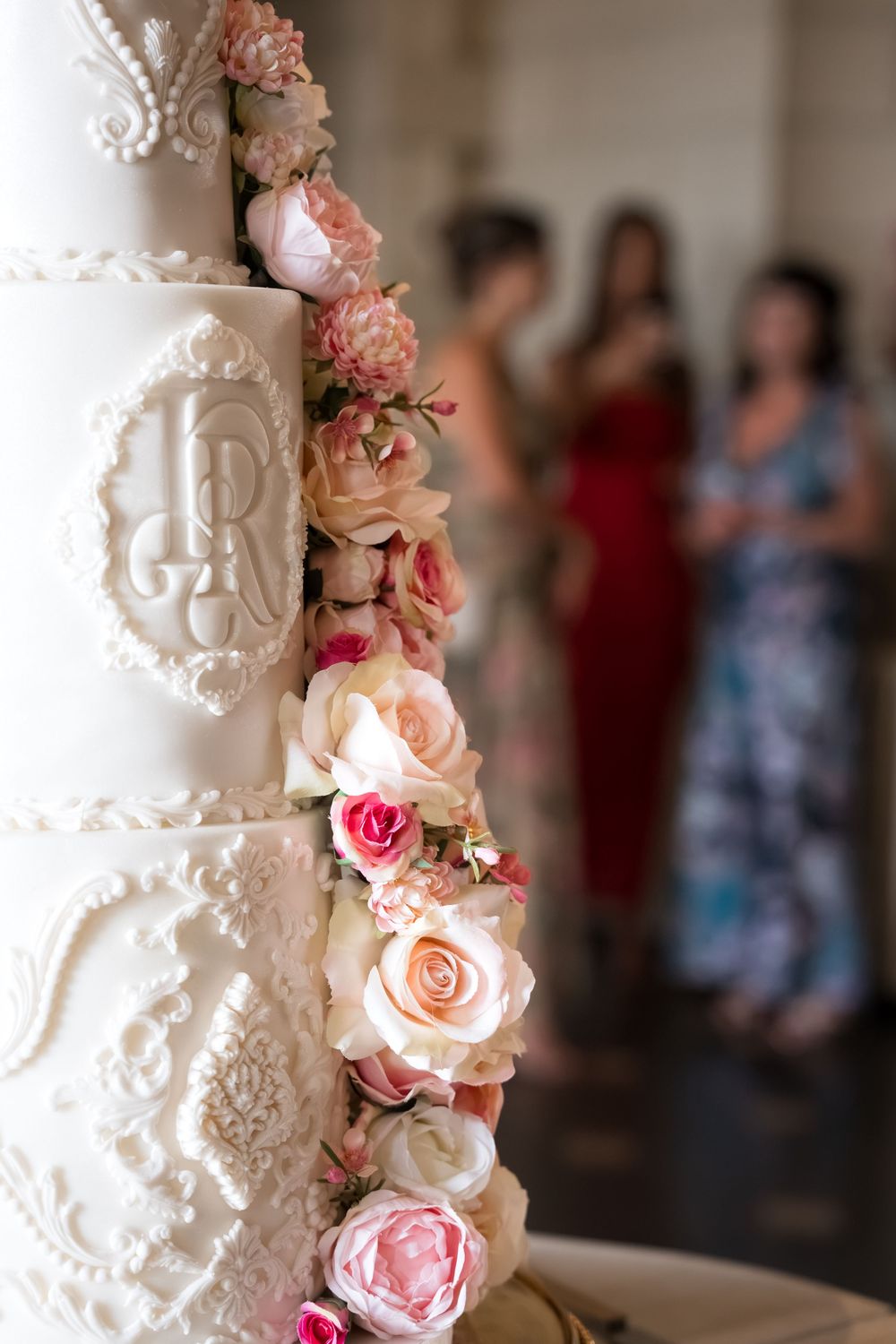 Elegant white wedding cake with pink roses and monogram detail, guests in colorful dresses blurred in background.