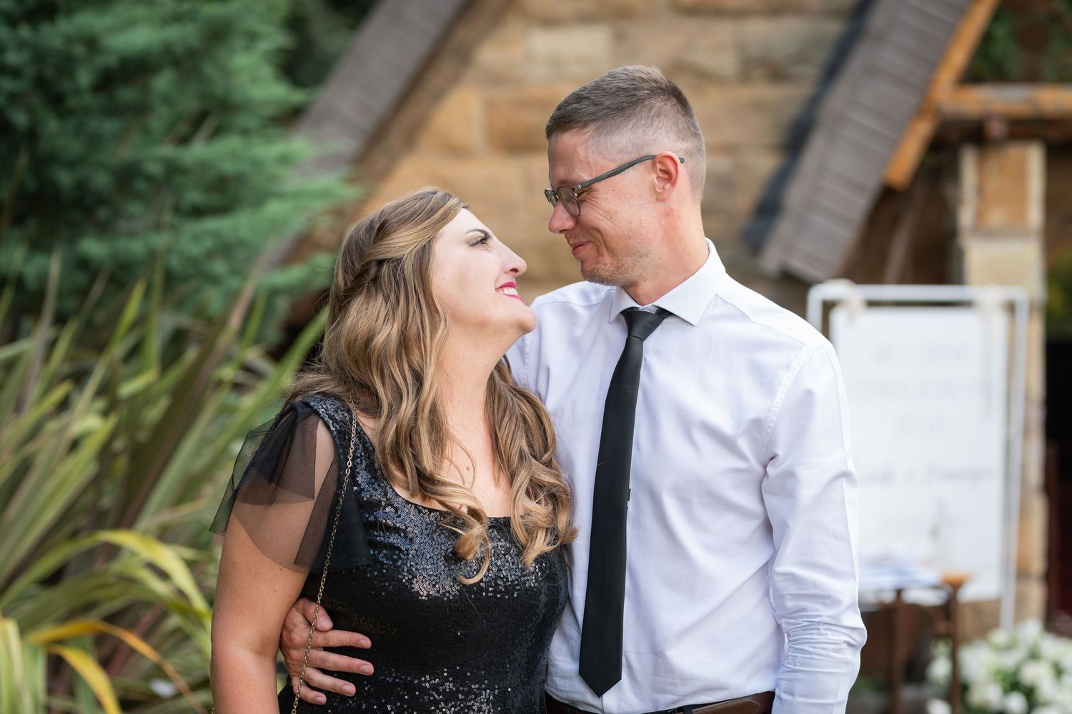 Couple in formal attire smiling at each other outdoors, woman in black dress, man in white shirt and tie.