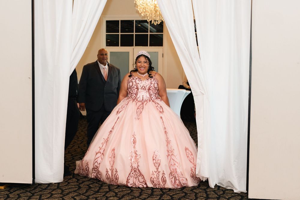 Young woman in pink ballgown with rose gold embroidery makes grand entrance at quinceañera celebration.