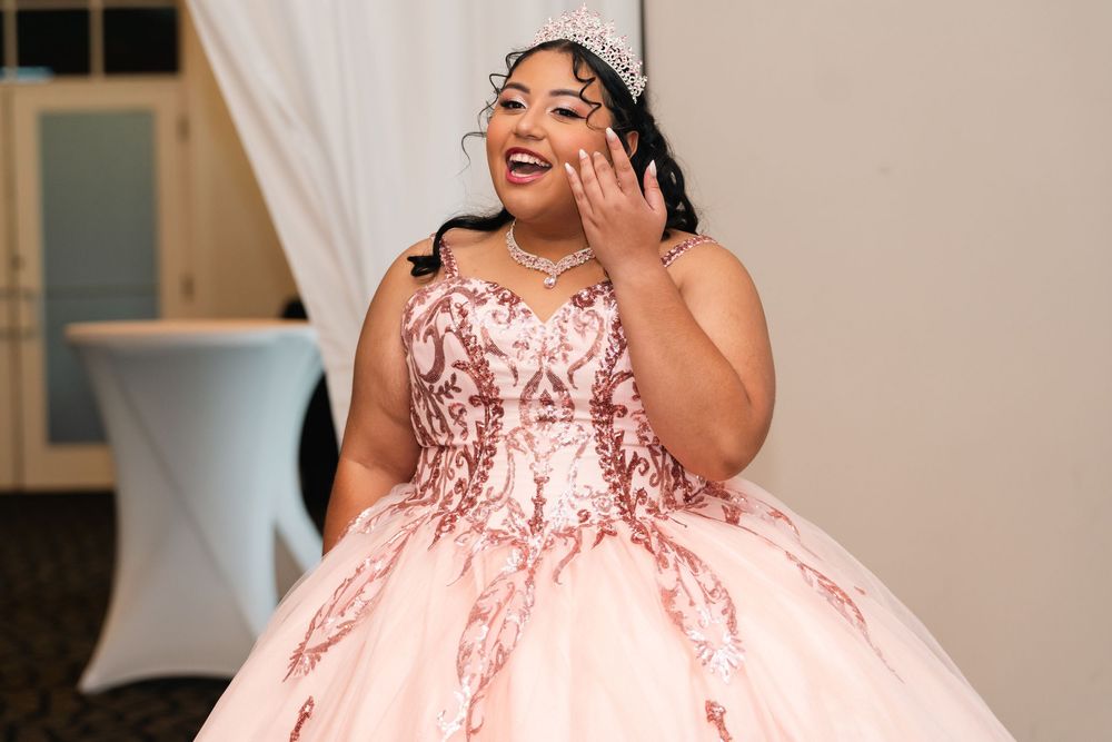 Young woman in pink quinceañera ball gown with rose gold sequins and tiara, smiling and waving at celebration.