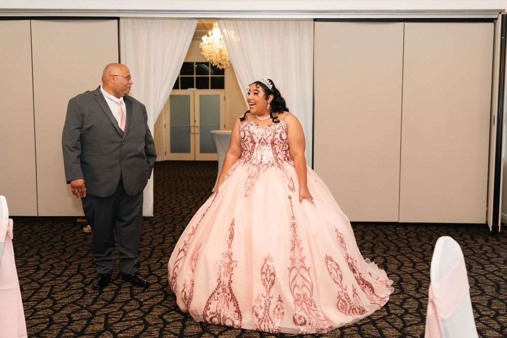 A young woman in a pink ball gown quinceañera dress stands with a man in a gray suit at an elegant event venue.