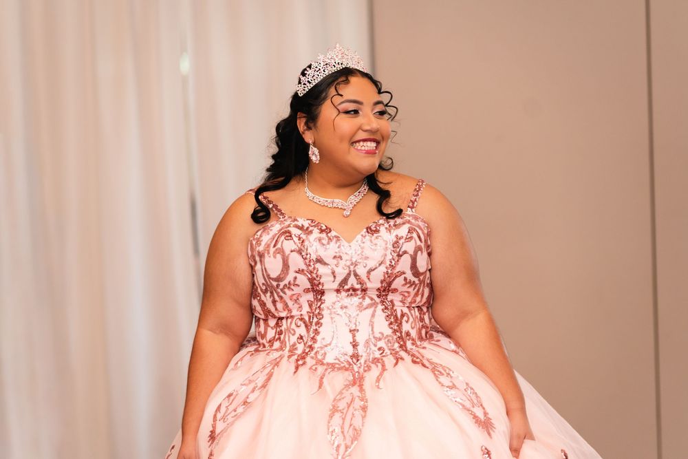 Young woman in pink quinceañera ball gown with tiara and jewelry smiling at formal celebration event.