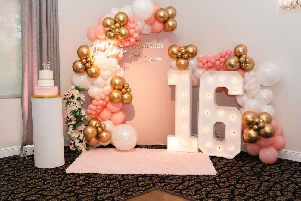 Pink and gold sweet 16 party backdrop with balloon garland, marquee number 16, and tiered cake display.