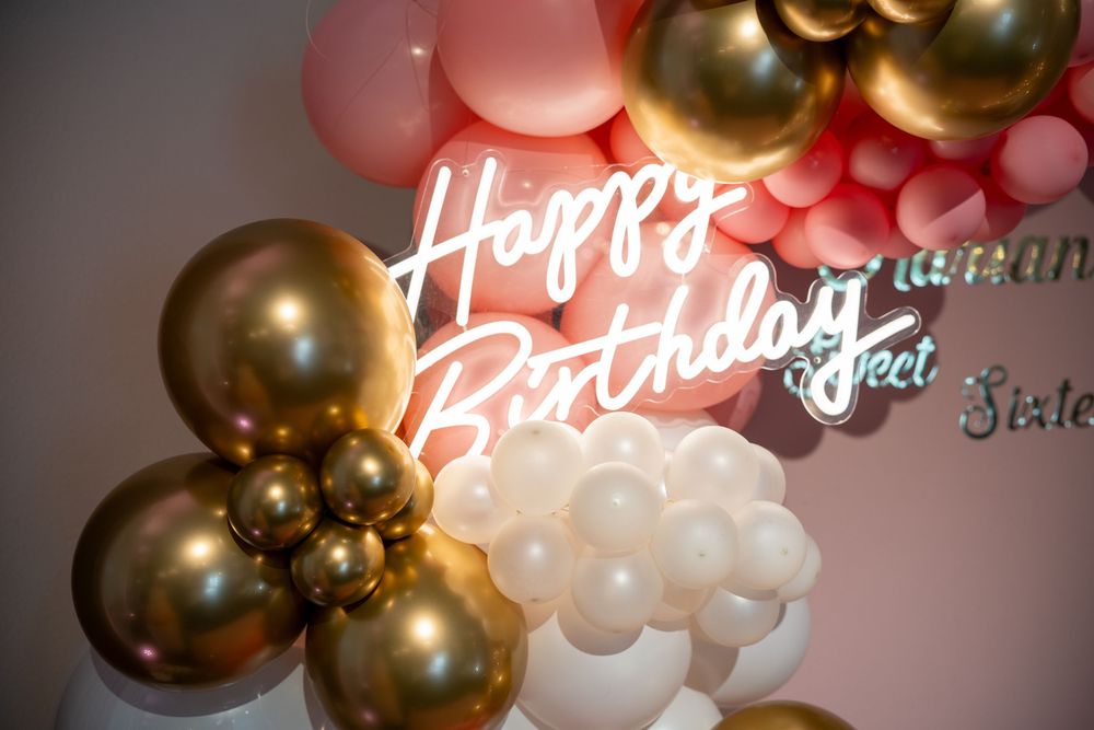 Neon Happy Birthday sign surrounded by gold, pink, and white balloon garland party decoration.